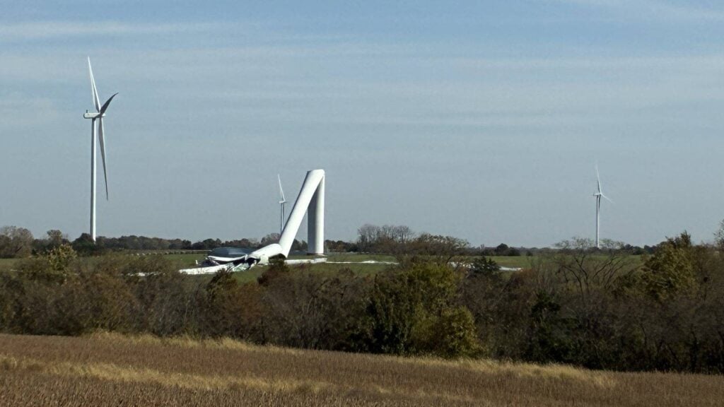 Wide view of the fifth collapsed turbine at High Prairie wind farm beyond a harvested field and tree line